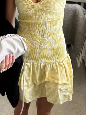 Gul rutig kortklänning från Zara - Supersöt gul och vit rutig kortklänning från Zara med smockad midja, volangkant och v-ringad byst. Perfekt för soliga dagar och har en feminin, figurnära passform. Tillverkad i mjuk bomull som känns skön mot huden.