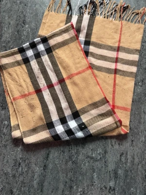 Beige halsduk med rutigt mönster - Snygg beige halsduk med klassiskt rutigt mönster i svart, vitt och rött. Halsduken har fransar i ändarna och är tillverkad i ett mjukt tyg som känns skönt mot huden. Perfekt accessoar för att lyfta din outfit.