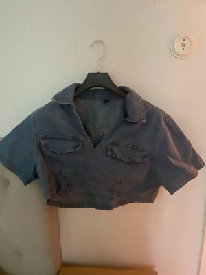 Blå croppad denimskjorta - Säljer en croppad blå skjorttopp i denim-look med korta ärmar och två bröstfickor. Toppen har krage och v-ringning, perfekt för en trendig och avslappnad stil.