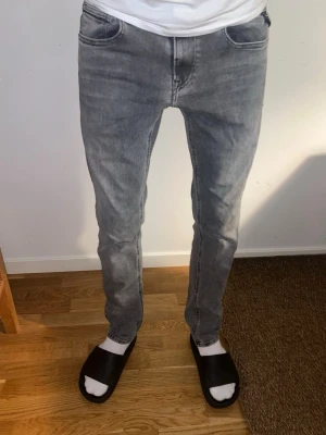 Gråa Replay Grover Jeans - Säljer ett par grå tvättade jeans med klassisk femficksmodell och raka ben. Jeansen har snygga slitningar och kontrastsömmar för en cool look. Perfekta till sneakers eller boots och passar till många olika stilar.
