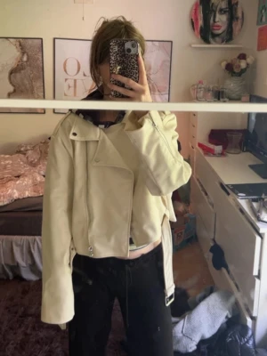 Vit croppad skinnjacka med bälte - Snygg vit skinnjacka i croppad modell med bred krage, dragkedja och bälte i midjan. Jackan har silverfärgade detaljer och dragkedjor vid fickorna. Perfekt för dig som vill ha en trendig och stilren look.