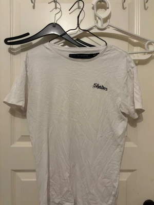 Vit t-shirt med Shades-tryck - Vit t-shirt med rund hals och korta ärmar. På bröstet finns ett litet svart tryck med texten 'Shades'. Enkel och clean design som passar till det mesta. Tillverkad i mjuk bomull för skön känsla.