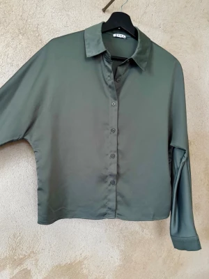 Grön skjorta från Wera - Stilren grön skjorta från Wera med klassisk krage och knappar framtill. Skjortan har lång ärm och är tillverkad i ett mjukt, glansigt material som ger en lyxig känsla. Perfekt för dig som gillar minimalistisk och clean stil.