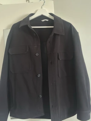 Svart overshirt med fickor - Stilren svart overshirt med klassisk krage och knäppning framtill. Två stora bröstfickor med lock och en mindre ficka på ena sidan. Overshirten har raka ärmar och en avslappnad passform. Perfekt att bära över en t-shirt för en cool lager-på-lager-look.