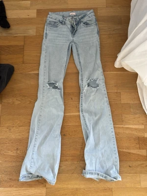 Ljusblå slitna bootcut jeans - Säljer ett par ljusblå bootcut jeans med slitningar på båda knäna och broderade detaljer i rosa på bakfickorna. Jeansen har klassisk femficksmodell och är tillverkade i denim. Perfekta för en avslappnad och trendig look. Slitage vid hälarna🩷