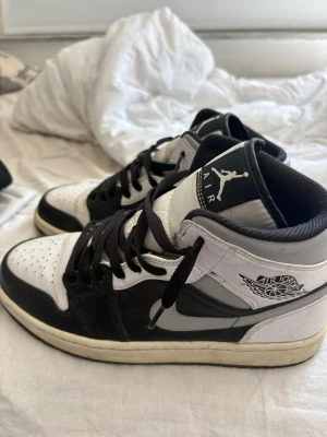 Air Jordan 1 Mid svart/vit - Säljer ett par Air Jordan 1 Mid sneakers i svart och vitt med klassisk vinge-logga på sidan och Jumpman på plösen. Skorna har svart snörning, vit tåbox och svart sula. Tillverkade i läder och textil för en clean och tidlös look.
