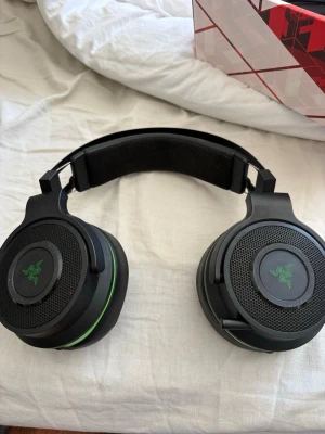Razer svarta gaminghörlurar - Säljer ett par svarta over-ear gaminghörlurar från Razer med gröna detaljer. De har vadderad bygel, stora öronkuddar i konstläder och inbyggda reglage för volym och mikrofon. Perfekta för gaming och långa sessions vid datorn.