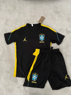 Svart  Brasilien träningsset  - Snyggt svart fotbollskit med Brasilien-emblem och gula detaljer. Setet består av en t-shirt och shorts, båda med gula och gröna inslag längs sidorna. T-shirten har BRASIL-tryck och ikonisk logga på ryggen. Perfekt för träning eller match.