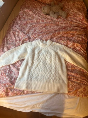 Vit stickad tröja med mönster - Mysig vit stickad tröja med klassiskt diamantmönster på framsidan. Tröjan har rund halsringning, långa ärmar och breda ribbade muddar vid ärmslut och nederkant. Perfekt för kalla dagar och enkel att matcha med olika outfits.