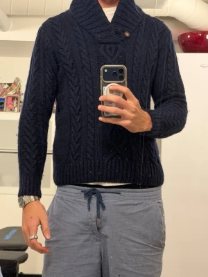 Mörkblå kabelstickad tröja från Gant - Snygg mörkblå kabelstickad tröja med sjalkrage och en knappdetalj vid halsen. Tröjan har långa ärmar och ribbade muddar vid ärmslut och nederkant. Perfekt för kyliga dagar och ger en klassisk look. Passar mera som S