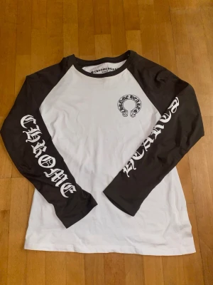Långärmad topp från Chrome Hearts - Säljer en långärmad topp från Chrome Hearts med svartvita kontraster. Ärmarna är svarta med vit text och kroppen är vit med en liten logga på bröstet. Perfekt för dig som gillar streetwear och vill sticka ut med en unik design.