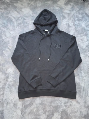 Svart hoodie med broderad logga - Snygg svart hoodie med huva och dragsnören. På bröstet finns en diskret broderad logga i svart. Hoodien har ribbade muddar vid ärmar och nederkant. Perfekt för en chill och stilren look.
