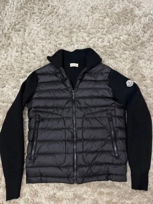 Svart pufferjacka med stickade ärmar - Snygg svart pufferjacka med quiltad front och stickade ärmar. Jackan har hög krage, två dragkedjefickor framtill och en Moncler-logga på vänster ärm. Materialmix med glansig nylon på kroppen och mjukt stickat tyg på ärmarna ger en modern look.