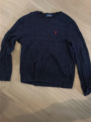 Mörkblå kabelstickad tröja Polo - Mörkblå kabelstickad tröja från Polo Ralph Lauren med rund halsringning och röd broderad logga på bröstet. Tröjan har ribbade muddar vid ärmslut och nederkant. Perfekt till jeans eller kjol för en klassisk look.