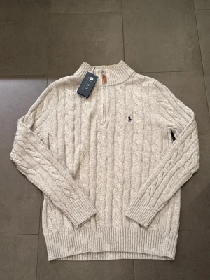 Ljusgrå kabelstickad tröja Polo Ralph Lauren - Snygg ljusgrå kabelstickad tröja  Polo Ralph Lauren med halv dragkedja och ribbade muddar. Tröjan har en liten broderad logga på bröstet och är gjord i mjukt material. Perfekt för dig som gillar klassisk och stilren look.