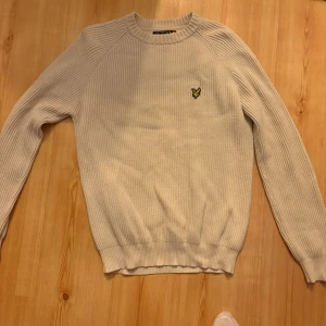 Beige stickad tröja Lyle & Scott - Säljer en beige stickad tröja från Lyle & Scott med rund halsringning och ribbade muddar. Tröjan har ett broderat gult logomärke på bröstet och är tillverkad i ett mjukt, ribbstickat material. Perfekt för lager på lager och enkel att matcha.