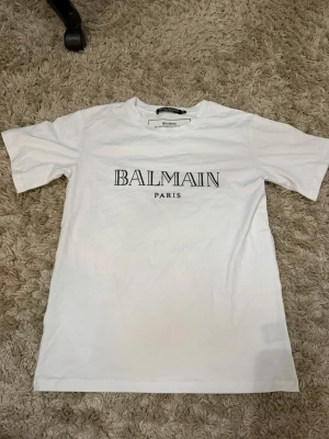 Vit Balmain Paris t-shirt XS - Snygg vit t-shirt från Balmain Paris med klassisk logga tryckt på bröstet. T-shirten har rund hals och korta ärmar. Tillverkad i mjukt bomullsmaterial, perfekt för en clean och stilren look.