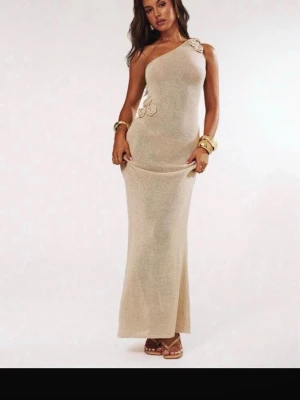 Beige oneshoulder långklänning - Elegant beige långklänning med oneshoulder-design och dekorativa blommor vid axeln och midjan. Klänningen har en smickrande, figurnära passform och är perfekt för dig som vill sticka ut med en stilren look.