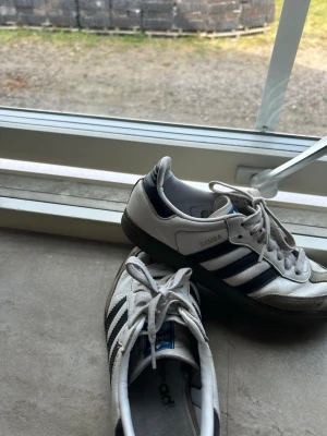 Adidas Samba vita och svarta sneakers - Klassiska Adidas Samba sneakers i vitt och svart med de ikoniska tre ränderna på sidan. Ovandelen är i läder med detaljer i mocka och yttersulan är i gummi. Skorna har blå Adidas-logga på plösen och Samba-tryck i guld på sidan.