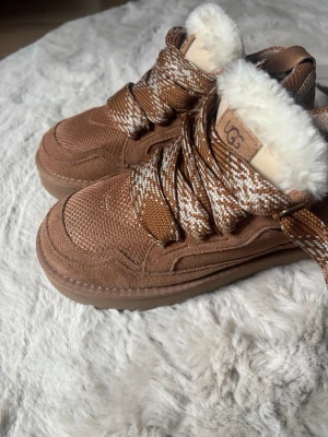 Bruna kängor med fuskpäls från UGG - Säljer ett par bruna kängor från UGG med grova snören i brunt och vitt. Kängorna har en fluffig vit fuskpäls runt öppningen och en robust sula. Materialet är en mix av mocka och textil, perfekta för kalla dagar. Aldrig använda då dem var försmå ( skulle säga att storleken är mer lik en 39/38 än 40 ) kommer med extra skosnören.