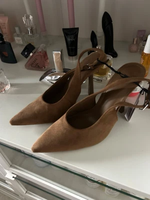 Bruna pumps med spetsig tå från Bershka - Snygga bruna pumps från Bershka med spetsig tå och öppen häl. Skorna har en smal klack och justerbar rem med spänne. Tillverkade i mockaimitation som ger en trendig och stilren look. Perfekta för att lyfta vilken outfit som helst.
