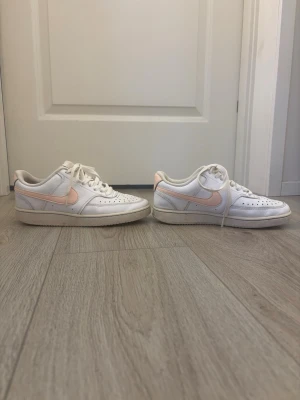 Nike sneakers med ljusrosa detalj - Snygga vita sneakers med ljusrosa swoosh på sidan. Skorna har klassisk låg profil, perforerad tå och vita skosnören. Tillverkade i läder med gummisula för bra grepp. Har dock ett litet släpp i sömnen på inre sidan av en av skorna syns inte när skon är på!