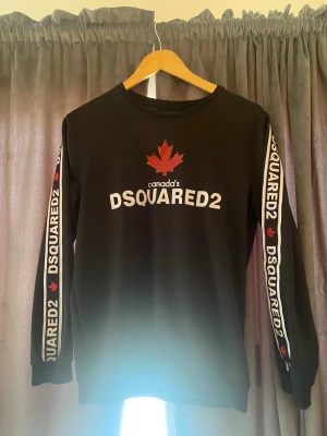 Svart långärmad tröja DSQUARED2 - Svart långärmad tröja från DSQUARED2 med vit logga och röd lönnlöv på bröstet. Texten 'DSQUARED2' och röda lönnlöv finns även längs båda ärmarna. Rund halsringning och avslappnad passform. Perfekt för dig som gillar streetwear och tydliga loggor.