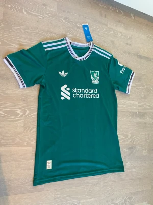 Grön Liverpool Adidas matchtröja - Snygg grön Liverpool FC matchtröja från Adidas med vita detaljer och klassiska tre ränder på axlarna.       Storlek L, Moch måtten finns på sista bilderna.                    Priset kan diskuteras och skickar inom 24 Tim☺️💨