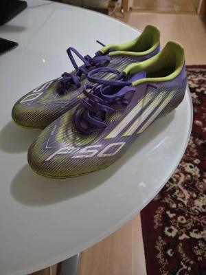 Lila Adidas F50 fotbollsskor league  - Säljer ett par lila Adidas F50 fotbollsskor med gula detaljer och vita ränder på sidan. Skorna har snörning och en låg profil, perfekt för snabba rörelser på planen. Yttermaterialet är syntetiskt och insidan är fodrad i gult för extra komfort.