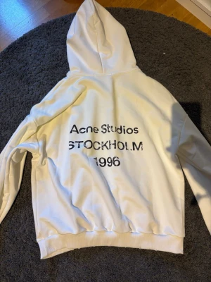 Vit hoodie Acne Studios Stockholm Medium - Säljer en vit hoodie från Acne Studios med trycket 'Acne Studios STOCKHOLM 1996' i svart på ryggen. Hoodien har klassisk passform, ribbade muddar och stor huva. Perfekt för dig som gillar stilrena och trendiga plagg med streetwear-vibe.