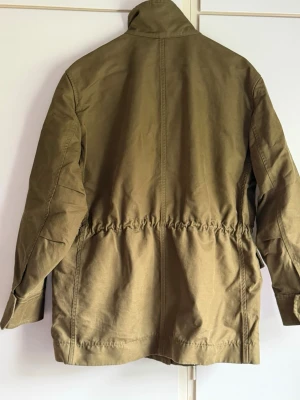 Olivgrön parkas med hög krage - Snygg olivgrön parkas med hög krage och fyra stora fickor framtill. Jackan har dragsko i midjan för en mer markerad siluett och är tillverkad i ett slitstarkt bomullsliknande material. Perfekt för dig som vill ha en stilren och funktionell jacka.