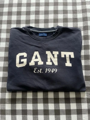 Mörkblå sweatshirt från GANT - Klassisk mörkblå sweatshirt från GANT med vit logga och texten 'Est. 1949' på bröstet. Tröjan har rund halsringning och mjuk insida, perfekt för en avslappnad och stilren look.