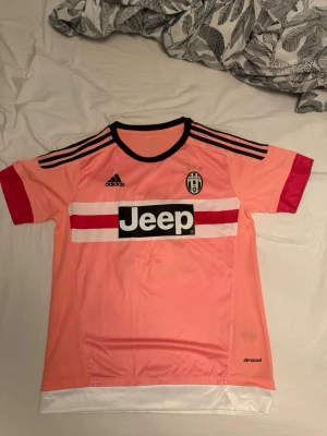 Juventus rosa matchtröja Dybala 21 - Juventus rosa matchtröja från Adidas med svarta detaljer, röda ärmslut och vita/röda ränder över bröstet. Jeep-logga på framsidan och Dybala 21 på ryggen. Tillverkad i lätt funktionsmaterial med climacool-teknologi. Perfekt för fotbollsträning eller som supporterplagg.