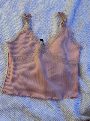 Rosa ribbad spetsbody från Divided - Söt ljusrosa ribbad body från Divided med tunna justerbara axelband och spetsdetaljer längs urringningen. Mjuk och stretchig kvalitet som sitter skönt mot kroppen. Perfekt att styla under en skjorta eller som den är.