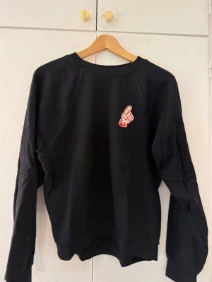 Svart sweatshirt med tryck - Svart sweatshirt med rund hals och ett litet tryck av en tecknad hand som pekar uppåt på bröstet. Tröjan har långa ärmar och ribbade muddar vid ärmslut och nederkant. Perfekt för en avslappnad och cool stil men som där med krumt så skulle säga ca S i storlek