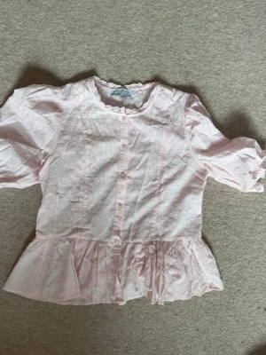 Ljusrosa blus med spets från Holly & Whyte - Söt ljusrosa blus från Holly & Whyte med spetsdetaljer längs knäppningen och runt halsen. Blusen har puffiga trekvartsärmar, volang nedtill och är tillverkad i ett tunt, broderat bomullstyg. Perfekt för en romantisk och feminin stil.