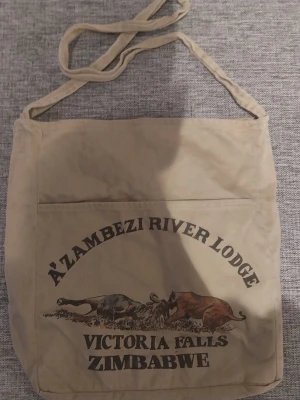 Beige shoppingväska med tryck - Säljer en beige shoppingväska i tyg med trycket 'A'Zambezi River Lodge Victoria Falls Zimbabwe' och motiv av två djur på framsidan. Väskan har en lång axelrem och en stor ytterficka. Perfekt för dig som gillar unika souvenirer och vill ha plats för mycket.