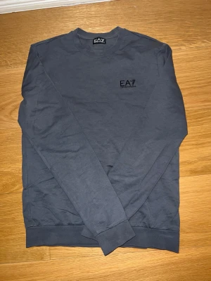 Mörkgrå sweatshirt från EA7 Armani - Snygg mörkgrå sweatshirt från EA7 Emporio Armani med rund halsringning och diskret logga på bröstet. Tröjan har långa ärmar och ribbade muddar vid ärmslut och nederkant. Perfekt för en clean och stilren look.