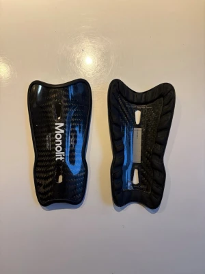 Monolit benskydd i kolfiber - Säljer ett par svarta benskydd från Monolit i kolfiber med ergonomisk form och vadderad insida. Lätta och smidiga för maximal rörelsefrihet på fotbollsplanen. Snygg design med kolfibermönster och vita detaljer.