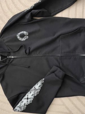 Svart Chrome Hearts hoodie med tryck - Säljer en svart hoodie från Chrome Hearts med vit logga på bröstet och stort tryck på ryggen. Ärmarna har detaljerade vita mönster och hoodien har dragkedja framtill samt fickor. Klassisk streetwear-look med huva och ribbade muddar.