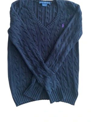 Polo Ralph Lauren Svart kabelstickad tröja  - Kabelstickad svart tröja från Polo Ralph Lauren med lila broderad logga på bröstet. V-ringad modell med ribbade muddar vid ärmslut och nederkant. Klassisk och tidlös design som passar till mycket.
