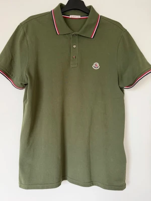 Olivgrön Moncler pikétröja - Snygg olivgrön pikétröja från Moncler med klassisk krage och knäppning framtill. Tröjan har vita och röda detaljer på krage och ärmslut samt Moncler-logga på bröstet. Perfekt för en stilren och avslappnad look. Kvitto och tags finns!