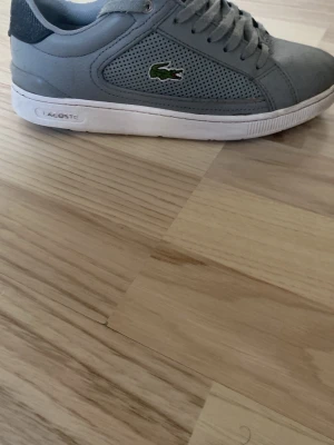 Gråa Lacoste sneakers  - Snygga gråa sneakers från Lacoste med klassisk låg profil och matchande grå skosnören. Skorna har diskret Lacoste-logga på plösen och är tillverkade i ett material som ser ut som mocka eller syntet. Perfekta för en clean och stilren look. Priset kan diskuteras😁