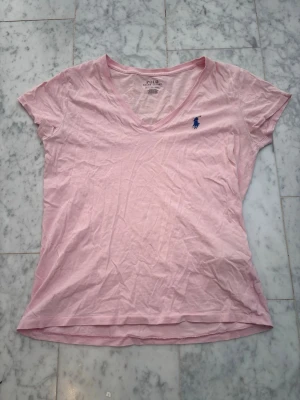 Ljusrosa v-ringad t-shirt Polo Ralph Lauren - Säljer en ljusrosa t-shirt från Polo Ralph Lauren med v-ringning och korta ärmar. T-shirten har den klassiska blå loggan broderad på bröstet och är gjord i mjuk bomull. Perfekt till jeans eller shorts för en avslappnad look.