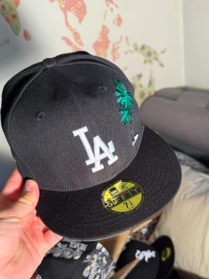 Svart LA Dodgers keps med palm - Snygg svart keps från New Era 59FIFTY med LA Dodgers-logga framtill och broderad palm i grönt. På sidan finns ett färgglatt 1988 World Series-märke. Klassisk flat brim och broderade ventilationshål. Perfekt för dig som gillar streetwear och sportig stil.