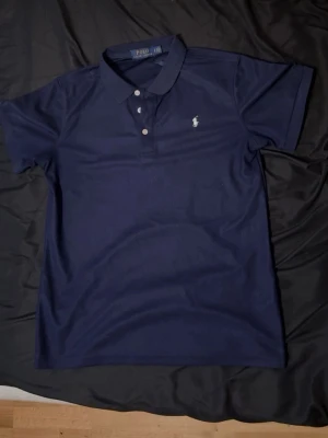 Marinblå pikétröja från Polo Ralph Lauren - Klassisk marinblå pikétröja från Polo Ralph Lauren med vit broderad logga på bröstet. Tröjan har korta ärmar, krage och knappar framtill. Perfekt till både jeans och shorts för en stilren look.