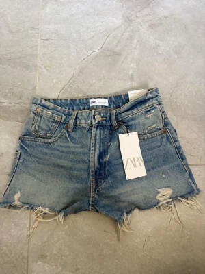 Blå högmidjade jeansshorts från Zara - Säljer ett par blå denimshorts från Zara med hög midja och fransiga, slitna kanter. Shortsen har klassisk femficksdesign, bälteshällor och knappgylf. Perfekta för en avslappnad och trendig sommarlook.