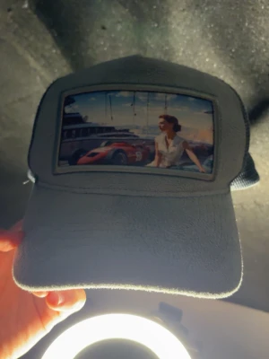 Grå meshkeps med retrotryck - Snygg grå keps med mesh på sidorna och baksidan för extra ventilation. Framtill finns ett retroinspirerat tryck med bil och kvinna vid en hamn. Perfekt accessoar för en cool och avslappnad stil.