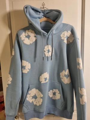 Ljusblå hoodie med molntryck - Säljer en ljusblå hoodie med vita molnliknande tryck över hela plagget. Hoodien har huva med dragsko och en stor magficka framtill. Perfekt för dig som gillar en chill och lekfull stil. Materialet känns mjukt och skönt mot huden. Ställ gärna frågor 