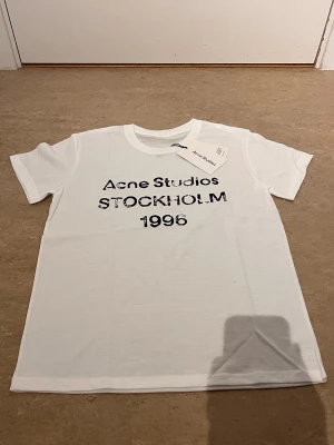 Vit Acne Studios t-shirt med tryck - Säljer en vit t-shirt från Acne Studios med svart tryck på bröstet där det står 'Acne Studios STOCKHOLM 1996'. Klassisk rund hals och korta ärmar. Perfekt basplagg med stilren design och skön passform.